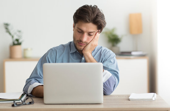 Tired Man Sitting At Laptop Touching Chin At Workplace