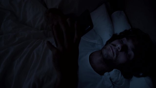Top view PAN shot of man lying awake in bed in dark and typing on mobile phone before going to sleep