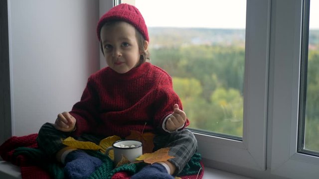 boy child in a red sweater and a knitted hat sits on a large white window
