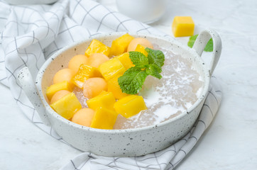 The thai dessert food modern style : The homemade sago in coconut milk with topping is mango cut into squares and cantaloupe ball in the white bowl and placed on tablecloth plaid on the marble table.