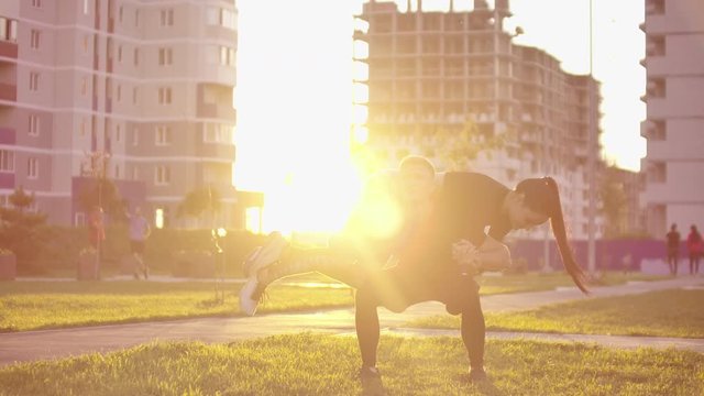A man squats with a girl sitting on his shoulders in a city Park against a backdrop of buildings at sunset in slow motion.