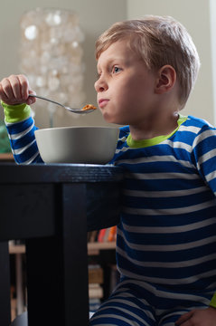 A cute little boy in striped pajamas eating cereal for breakfast at the dining room table