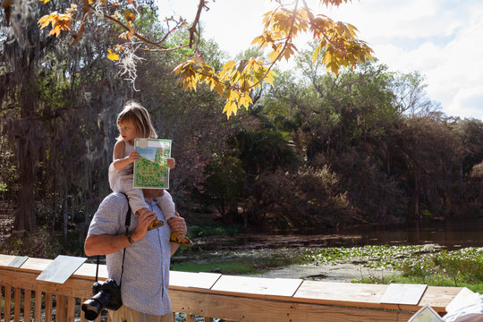 A little girl holding a map sitting on her dad's shoulders at a park 