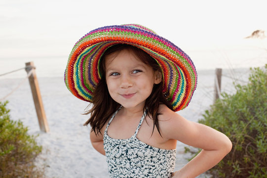 A cute little girl wearing a colorful straw hat smiling with dimples at the beach