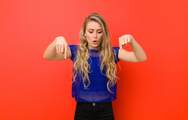 young blonde woman feeling shocked, open-mouthed and amazed, looking and pointing downwards in...