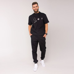 young stylish nurse male in black medical costume with stethoscope on his neck is standing with hands in pockets and smiling on the white wall background. medical fashion concept. free space