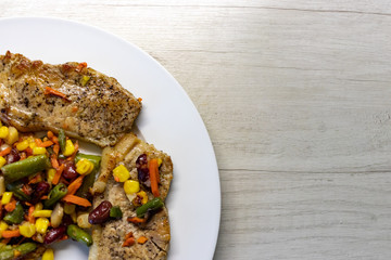 Mix of vegetable with fried meat on white plate of the light table. Mix of vegetable containing carrots, peas, beans and corn. Fried pork on a plate