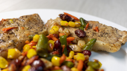 Mix of vegetable with fried meat on white plate of the light table. Mix of vegetable containing carrots, peas, beans and corn. Fried pork on a plate