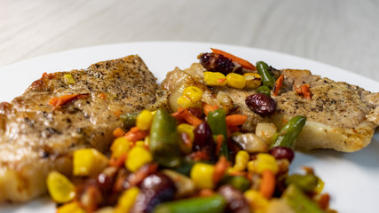 Mix of vegetable with fried meat on white plate of the light table. Mix of vegetable containing carrots, peas, beans and corn. Fried pork on a plate