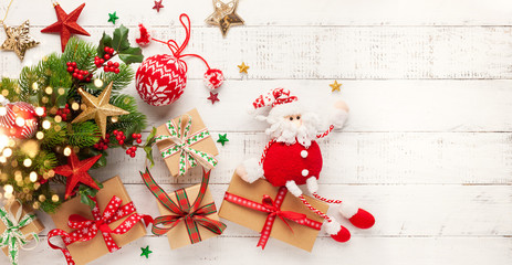 Christmas background with gift boxes, santa claus toy and branches of holly and fir on white wooden background. Winter festive concept. Flat lay, copy space.
