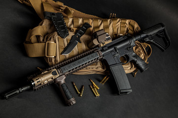 Modern automatic carbine with collimator sight on a dark background. Military backpack, knife and...