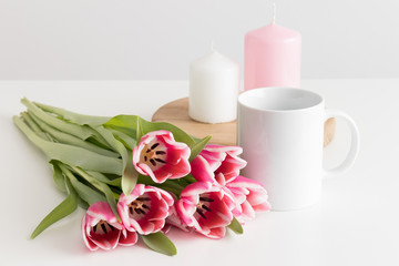 Bouquet of pink tulips, white mug and candles on a white table.