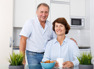Happy mature couple posing near kitchen table and drinking tea