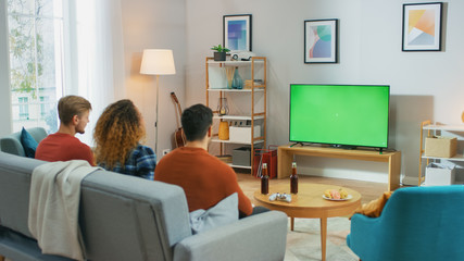 Three Friends Sitting on a Couch at Home, Watch Green Chroma Key Screen TV while Eating Snacks and Drinking Beverage. Young People Having Fun at Home.