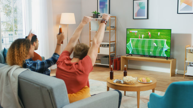 Three Sports Fans Sitting on a Couch in the Living Room Watch Important Soccer Match on TV, Cheering For their Team, Celebrates Doing YES Gesture after the GOAL Brings Victory to His Team.