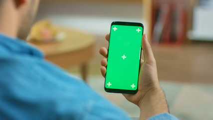 Man at Home Sitting on a Couch Holds Green Chroma Key Screen Touchscreen Smartphone. Browses Through Social Network, Reads News or Uses App.