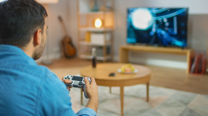 In the Living Room Man Sitting on a Couch Holds Controller Playing in a Console Video Game, 3D Action Shooter Gameplay Shown on TV Screen. © Gorodenkoff