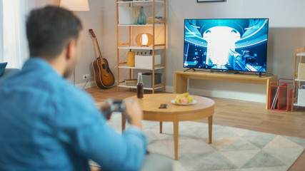 In the Living Room Man Sitting on a Couch Holds Controller Playing in a Console Video Game, 3D Action Shooter Gameplay Shown on TV Screen.