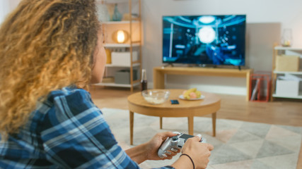 In the Living Room Girl Sitting on a Couch Holds Controller Playing in a Console Video Game, 3D Action Shooter Gameplay Shown on TV Screen.