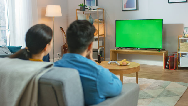 Happy Couple Sitting At Home Watching Green Chroma Key Screen Television, Relaxing On A Couch. Couple Room Watching Sports Game, News, Sitcom TV Show Or A Movie.