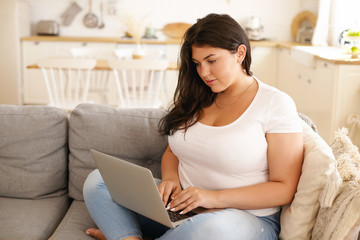 Adorable plus size student girl with loose black hair keyboarding on laptop sitting comfortably on...