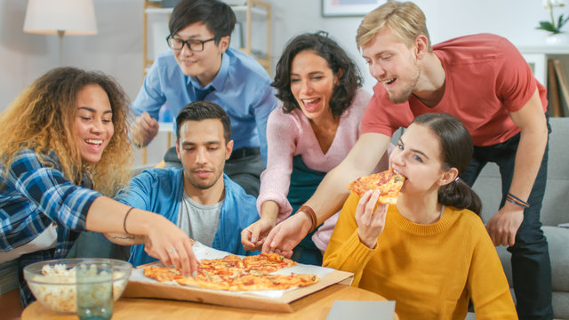 At Home Diverse Group Friends Watching TV Together, They Share Gigantic Pizza, Eating Tasty Pie Pieces. Guys And Girls Watching Comedy Sitcom Or A Movie, Laughing And Having Fun Together.