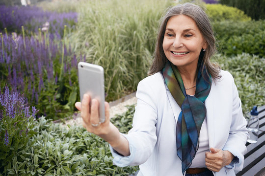 Outdoor picture of joyful positive stylish female with silk scarf around her neck posing against amazing background, sitting on bench, surrounded with green bushes and violet flowers, taking selfie