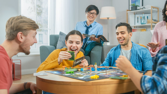 Diverse Group of Guys and Girls Playing in a Strategic Board Game with Cards and Dice. Man Throws Dice Impressively. Cozy Living Room in a Daytime