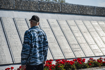 An elderly man stands in front of a memorial wall with the names of the victims of the second world...