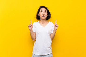 Asian young woman over isolated yellow wall surprised and pointing up