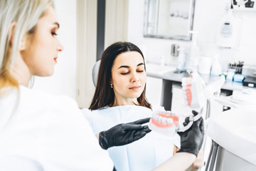 Pretty female dentist explaining way of treatment on the denture for the patient in the clinic.