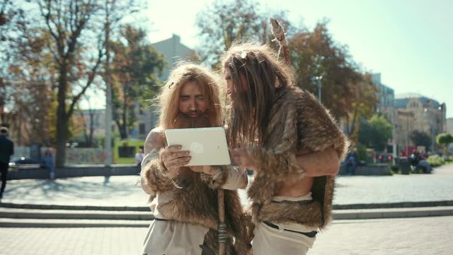 Modern-day neanderthals using digital tablet in present city. Archaic smart cavemen browsing internet content outdoor. Human adaptation concept.