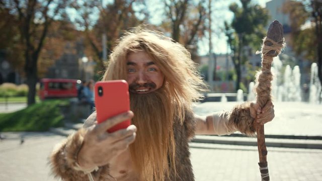 Portrait of wildman hunter-gatherer holding modern device making scary fun selfies outdoor. Primeval hipster using smartphone in modern city center.
