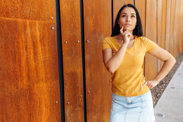 Thinking brunette young woman with bright smile dressed in casual clothes in front of rusty wall.