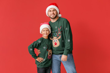 Fototapeta premium Happy man with his son in Christmas sweaters and Santa hats on color background
