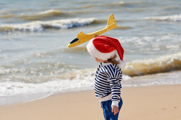 Little girl in Santa hat playing with toy airplane on sunny tropical beach. Christmas and new year vacation travel concept