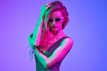 Fashionable glamour woman with Trendy wavy neon hairstyle. Party night club vibes. Excited shapely sexy girl in Stylish outfit, sunglasses. Bright pink purple light. Art fashion neon color.