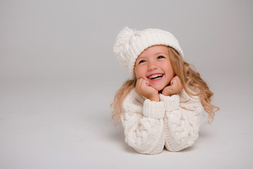 Winter clothes. Portrait of a little curly-haired girl in a knitted white winter hat. little blonde girl in white knitted hat and sweater smiling light background isolate, space for text