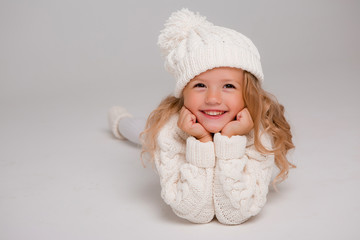 Winter clothes. Portrait of a little curly-haired girl in a knitted white winter hat. little blonde girl in white knitted hat and sweater smiling light background isolate, space for text