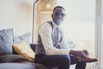 Posh attractive man in sunglasses is dressing up his leather brown shoes while sitting on sofa.