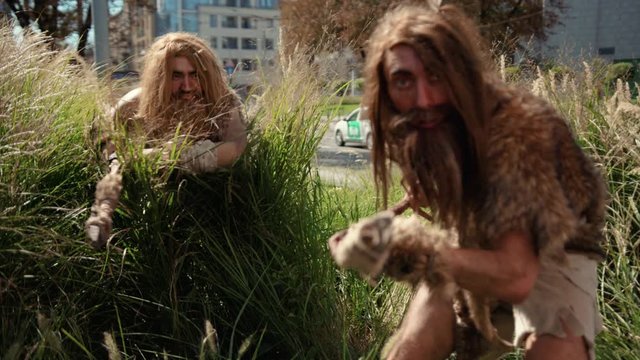Scared tribesmen of hunter-gatherers walking slowly through bushes on sunny day. Prehistoric cave people hunting in modern cityscape background.