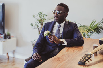 Handsome elegant groom in sunglasses is sitting next to the table with flower in his hand.