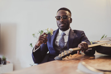 Handsome elegant groom in sunglasses is sitting next to the table with flower in his hand.
