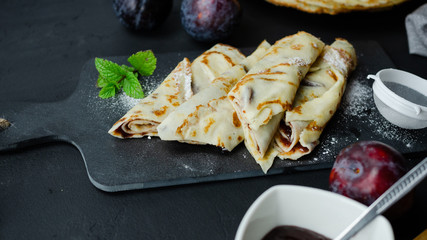 Naleśniki z powidłami śliwkowymi leżące na czarnej kamiennej desce. Ciemne tło.