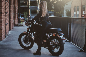 Obraz na płótnie Canvas Stylish female biker is sitting at her black bike while posing for a photoshoot on the street.