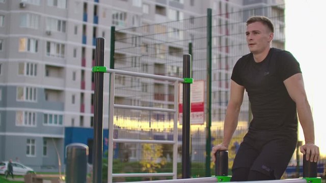 A man performs a leg lift on parallel bars in slow motion against the background of buildings in a city Park