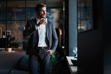 Young handsome businessman stand and hold glass of whiskey in his own office.