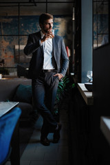 Young handsome businessman stand and hold glass of whiskey in his own office.