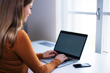 Young female persong typing on laptop on a white desk by the window