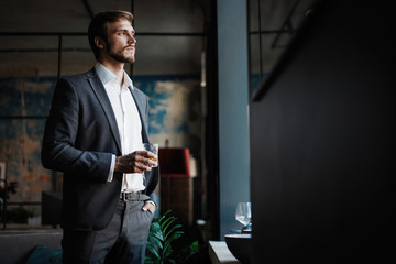 Young handsome businessman stand and hold glass of whiskey in his own office.